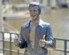 Life-size silver statue of a smiling man in a jacket giving thumbs up by a riverside railing.