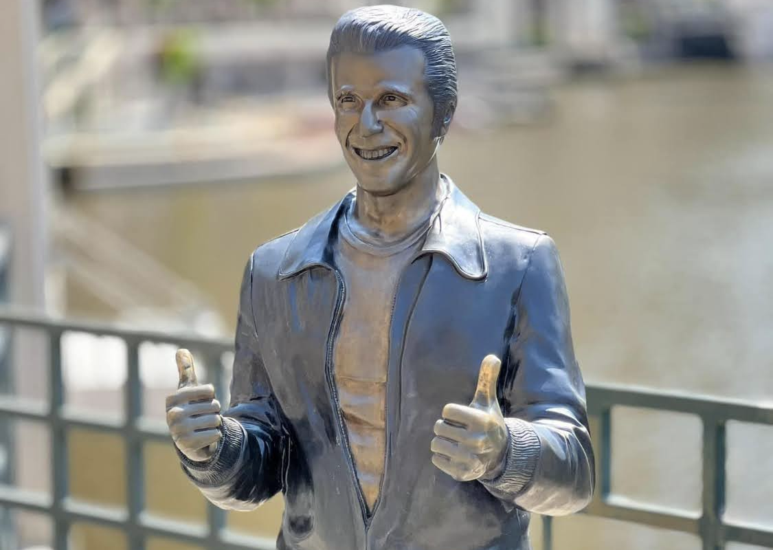 Life-size silver statue of a smiling man in a jacket giving thumbs up by a riverside railing.