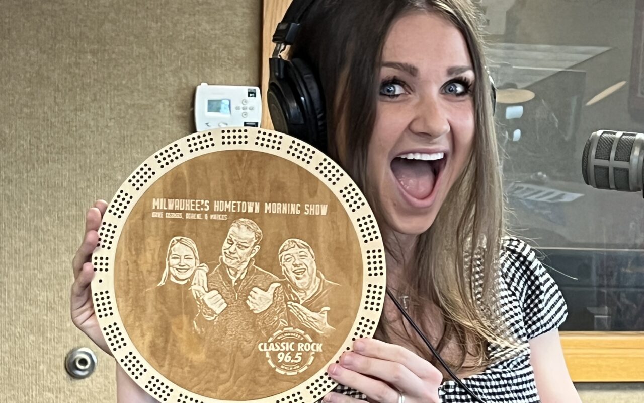 Smiling woman in a radio studio wearing headphones, holding a circular wooden plaque for Milwaukee's Hometown Morning Show.