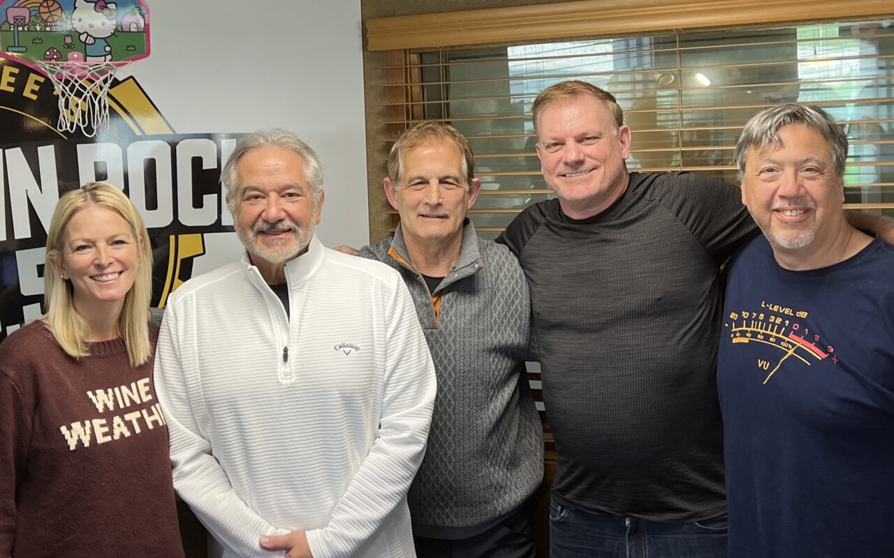 Five adults posing in a studio, smiling, with a sports-themed backdrop and blinds behind them.