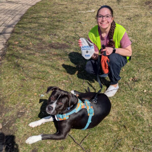A smiling woman kneels on grass with a black-and-white dog wearing a bright teal harness, outdoors in a park setting.