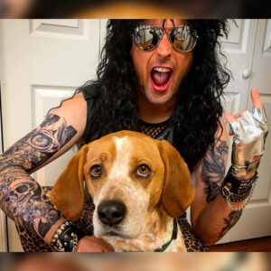 Person with long black hair, sunglasses, and full-arm tattoos poses with a beagle; wearing a metallic glove and leopard-print outfit.