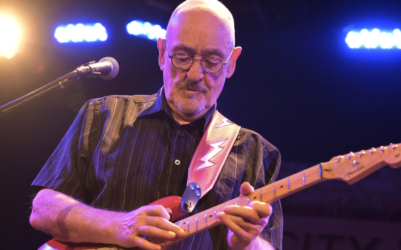 Older man with glasses playing a red electric guitar on stage, pink strap, singing into a microphone nearby