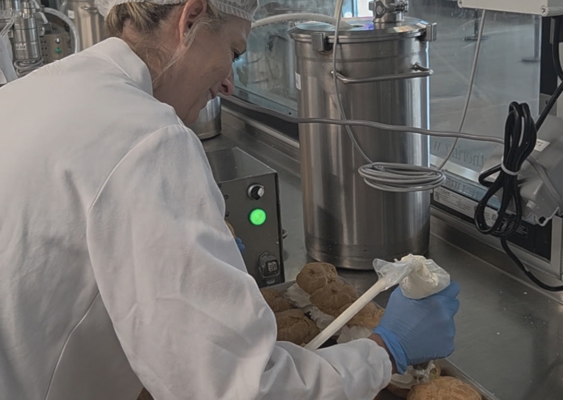 Person in a white lab coat, hairnet, and blue gloves piping cream onto pastries on a stainless production line.