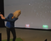 Woman in a blue shirt swinging a golf club in an indoor simulator; projection shows red WAIT and green HIT buttons on the screen behind her.