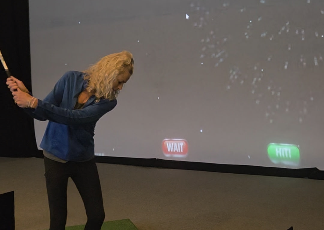 Woman in a blue shirt swinging a golf club in an indoor simulator; projection shows red WAIT and green HIT buttons on the screen behind her.