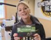 Smiling woman in a radio studio holds a green-dotted package of 'It's a Big Dill' pickle-flavored fudge toward the camera.