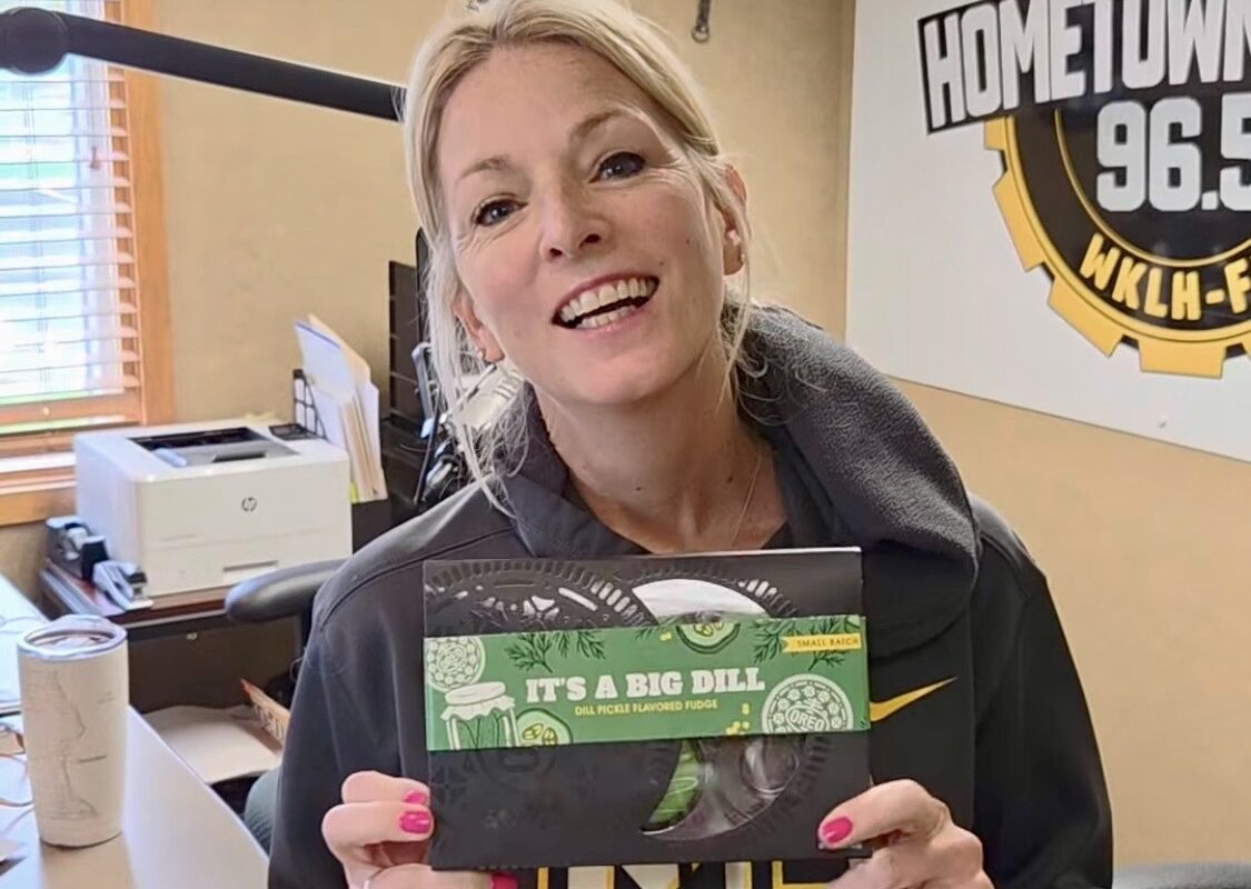 Smiling woman in a radio studio holds a green-dotted package of 'It's a Big Dill' pickle-flavored fudge toward the camera.