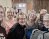 Six women smiling for a group photo indoors, standing close together near a wooden wall and doorway.