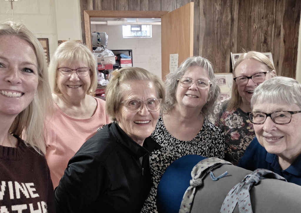 Six women smiling for a group photo indoors, standing close together near a wooden wall and doorway.
