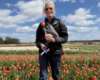 Smiling woman in a black quilted jacket and sunglasses holds a red tulip in a colorful tulip field on a sunny day.