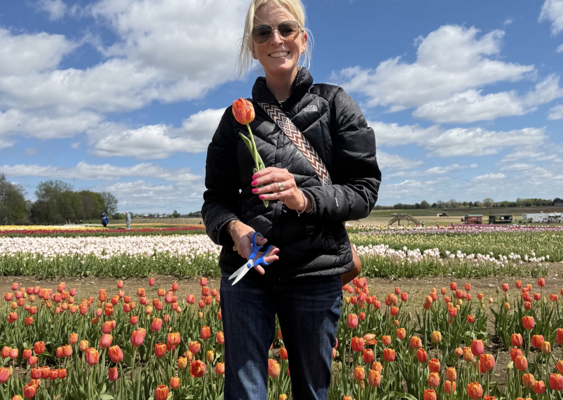 Smiling woman in a black quilted jacket and sunglasses holds a red tulip in a colorful tulip field on a sunny day.