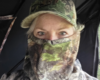 Person in camouflage cap and face covering, with only the eyes visible, looking at the camera in a camo-filled setting.