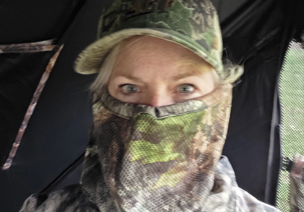 Person in camouflage cap and face covering, with only the eyes visible, looking at the camera in a camo-filled setting.