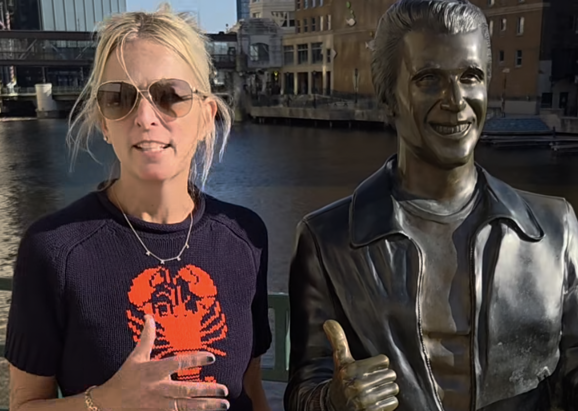 Woman with sunglasses standing beside a shiny silver statue that is giving a thumbs up, by a waterfront in a city.