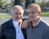 Two smiling men with arms around each other posing in a sunny parking lot on a casual day.