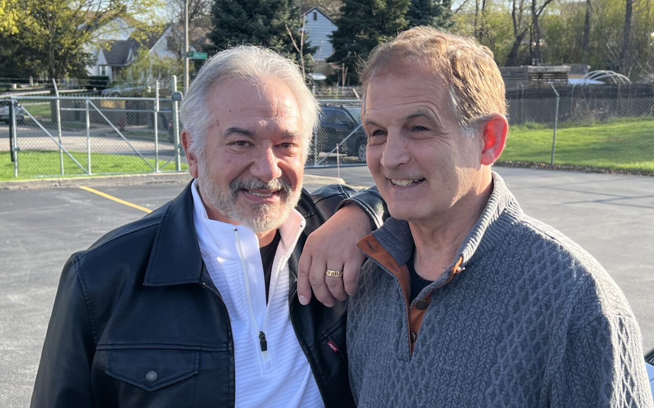 Two smiling men with arms around each other posing in a sunny parking lot on a casual day.