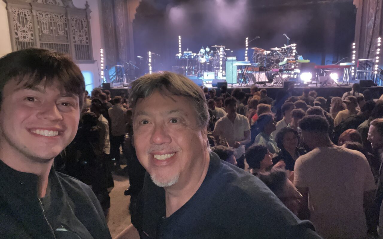 Two men smile for a selfie in a crowded concert venue with a stage and instruments in the background.
