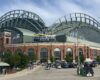 Exterior view of American Family Field, a brick stadium with a distinctive curved green roof and crowds outside on a sunny day.