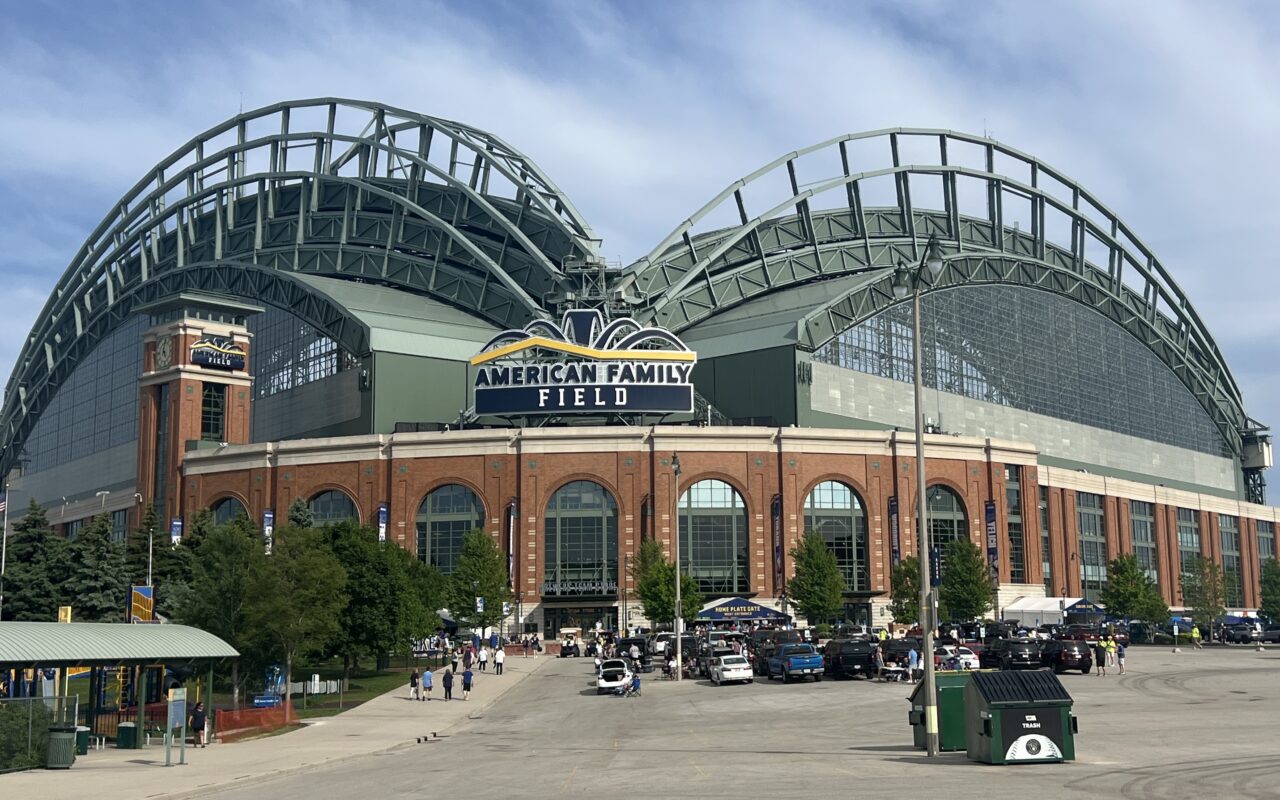 Exterior view of American Family Field, a brick stadium with a distinctive curved green roof and crowds outside on a sunny day.