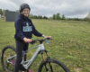 Smiling woman in a helmet standing beside a gray full-suspension mountain bike in a grassy field with trees in the background.