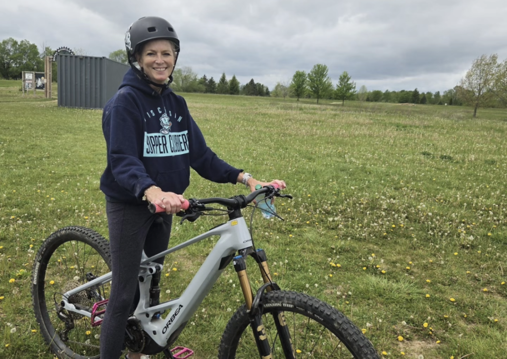 Smiling woman in a helmet standing beside a gray full-suspension mountain bike in a grassy field with trees in the background.