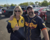 Two smiling adults pose together in a parking lot; the woman wears a yellow-blue jacket and sunglasses, the man wears a dark jersey with 'Fat Dog Club' logo and a cap, both arm-in-arm and smiling at the camera.