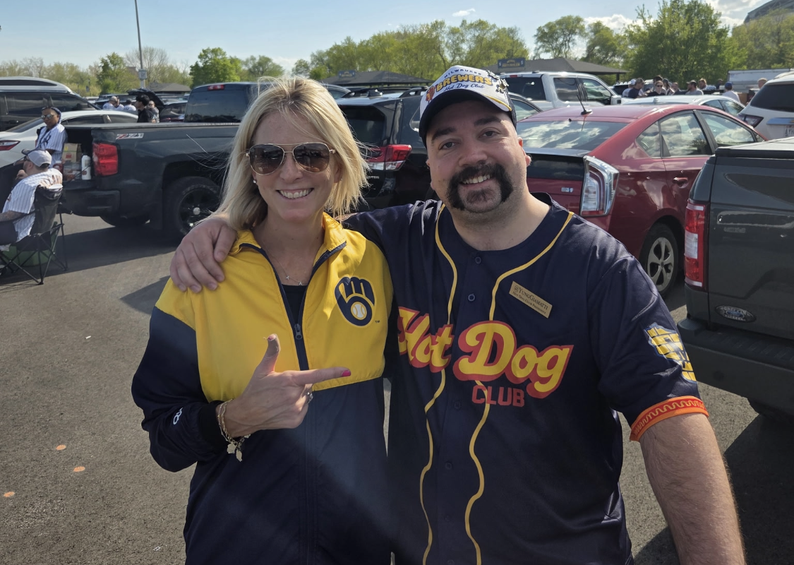 Two smiling adults pose together in a parking lot; the woman wears a yellow-blue jacket and sunglasses, the man wears a dark jersey with 'Fat Dog Club' logo and a cap, both arm-in-arm and smiling at the camera.