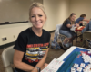 Smiling woman seated at a game table with tile pieces and score sheets, participating in a group activity.