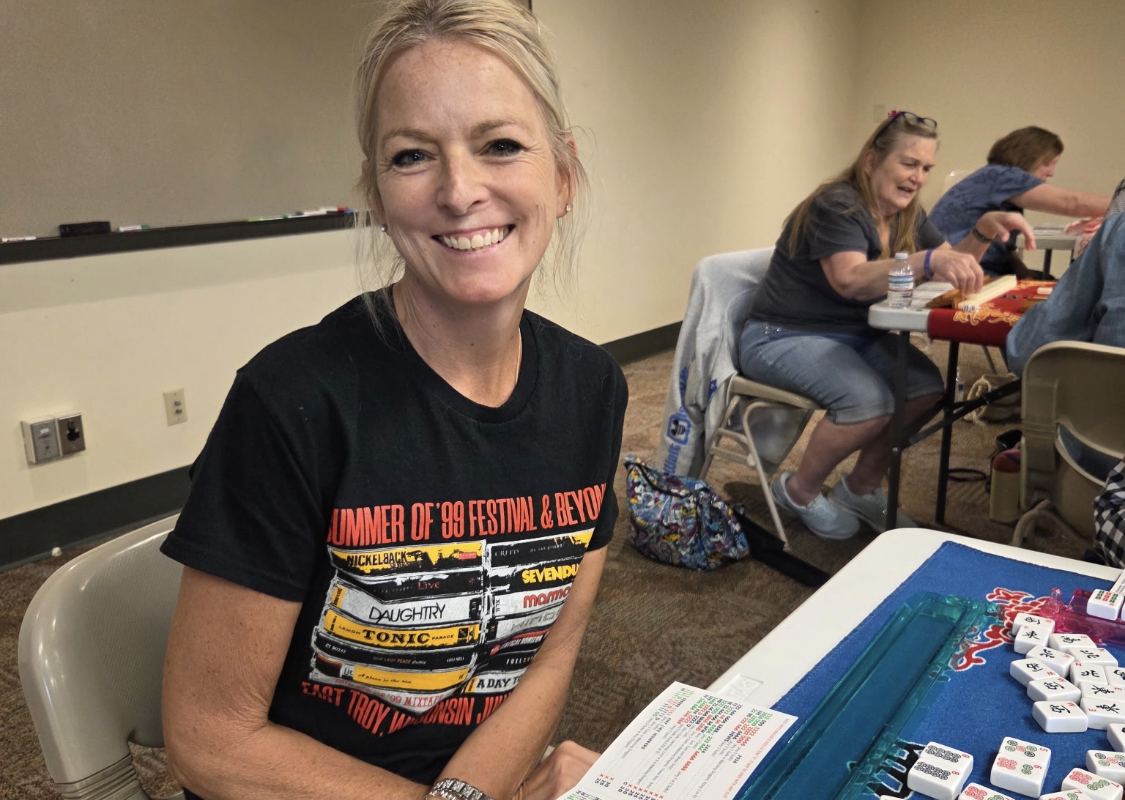 Smiling woman seated at a game table with tile pieces and score sheets, participating in a group activity.