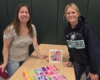 Two women stand at a table, smiling, with colorful puzzle pieces arranged on the surface and a puzzle box between them in the background.