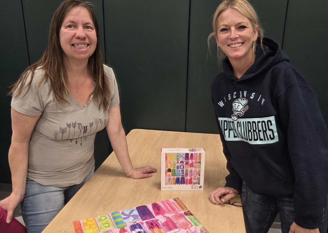 Two women stand at a table, smiling, with colorful puzzle pieces arranged on the surface and a puzzle box between them in the background.