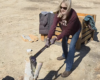 Woman wearing sunglasses splits a log with an axe in a dusty outdoor yard.