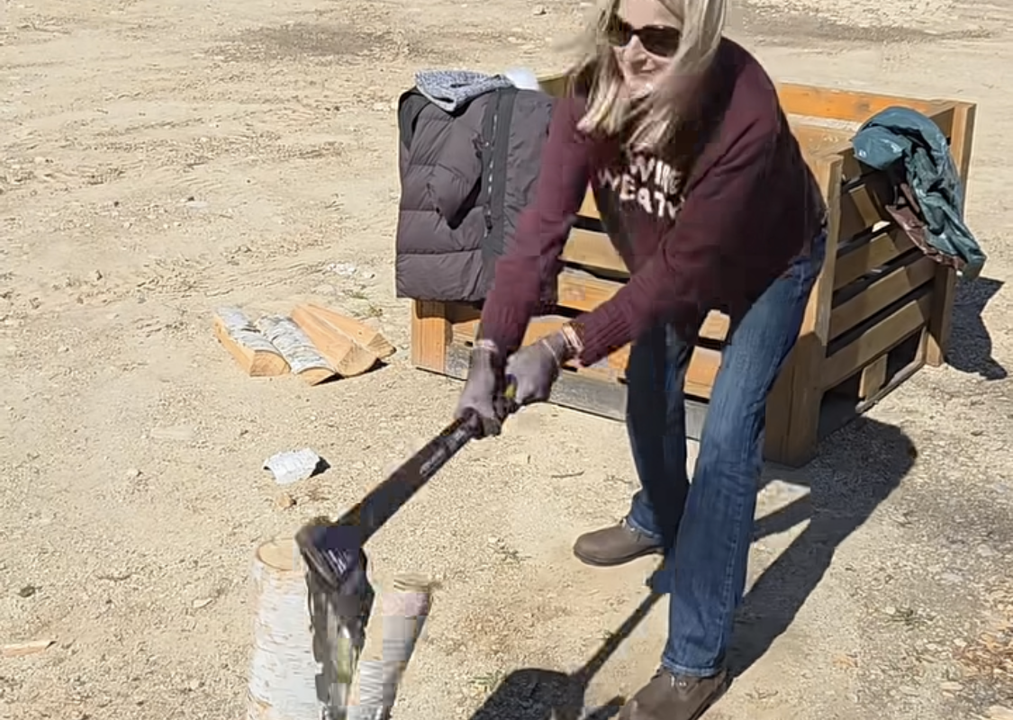 Woman wearing sunglasses splits a log with an axe in a dusty outdoor yard.