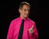 Man in a bright pink shirt and black tie pointing at the camera against a dark background.