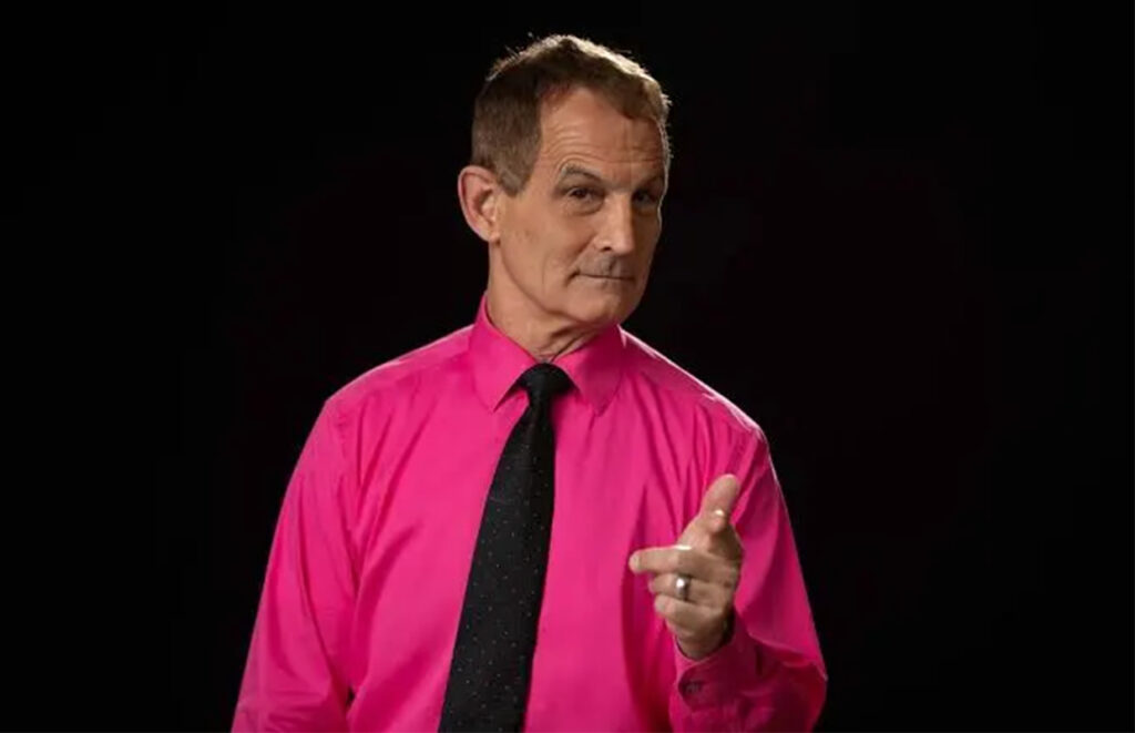 Man in a bright pink shirt and black tie pointing at the camera against a dark background.