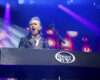 Male lead keyboardist sings into a mic on a lit stage behind a large keyboard with a Styx logo, colorful spotlights ablaze around him.