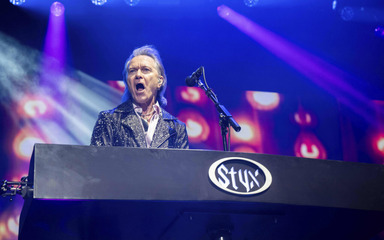 Male lead keyboardist sings into a mic on a lit stage behind a large keyboard with a Styx logo, colorful spotlights ablaze around him.