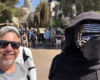 Smiling man with sunglasses takes a selfie with a person in a black hood and masked Kylo Ren costume at a sunny outdoor event; a group of people in the background.
