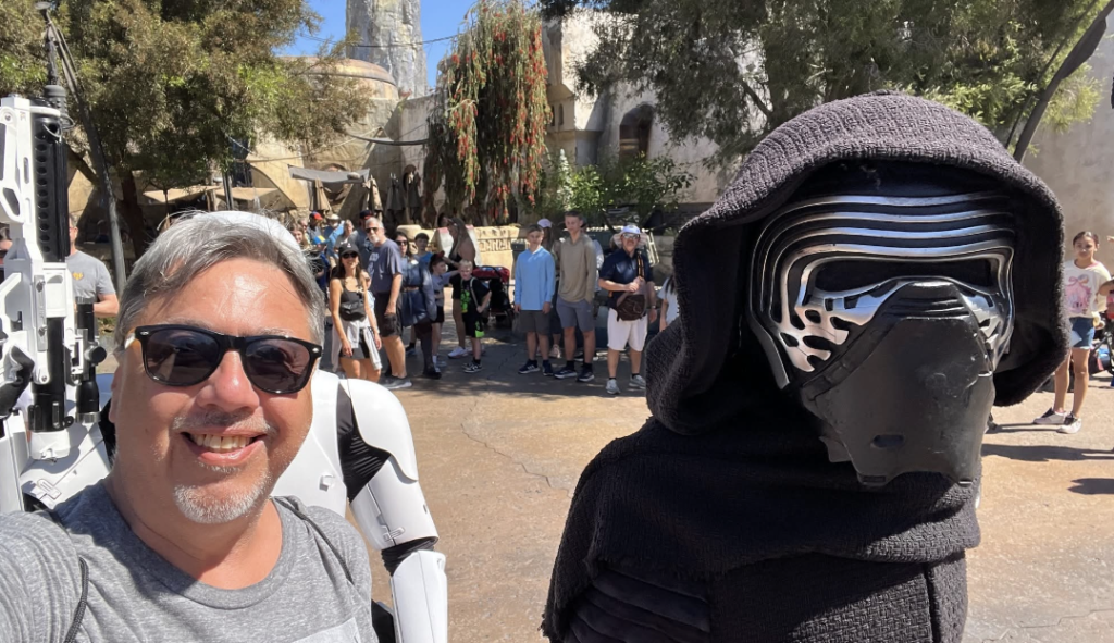 Smiling man with sunglasses takes a selfie with a person in a black hood and masked Kylo Ren costume at a sunny outdoor event; a group of people in the background.