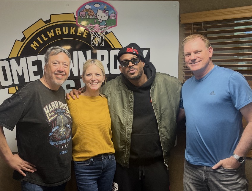 Four adults pose together indoors in front of a Milwaukee-themed backdrop, smiling at the camera.