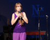Female comedian in a purple sequined sleeveless top and pants, holding a microphone on stage with stage equipment nearby.