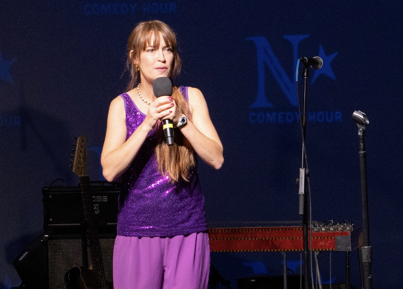 Female comedian in a purple sequined sleeveless top and pants, holding a microphone on stage with stage equipment nearby.
