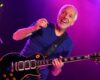 Guitarist with short white hair smiling while playing a dark electric guitar on stage under purple lights.] ,Oops