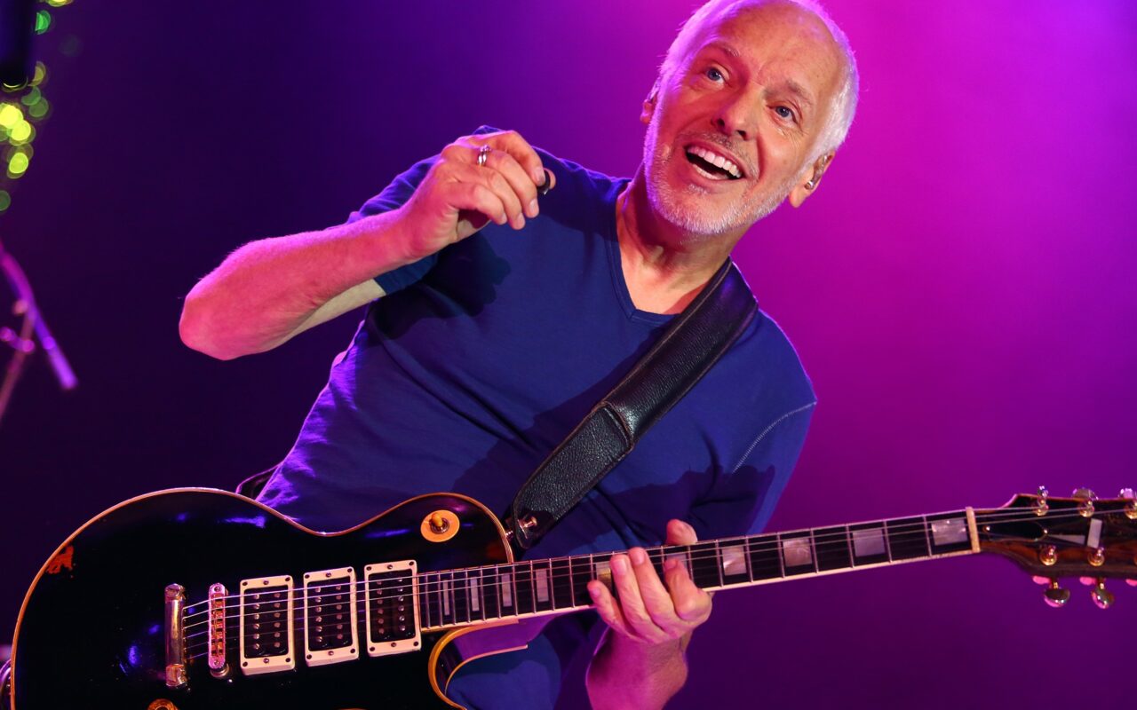 Guitarist with short white hair smiling while playing a dark electric guitar on stage under purple lights.] ,Oops