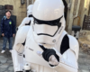 A white Stormtrooper in armor points toward the camera, standing in a themed outdoor area with a person in the background.