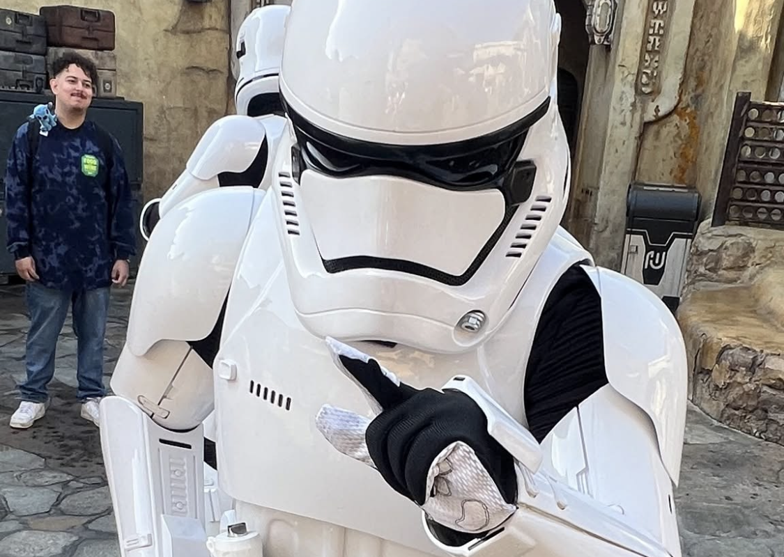 A white Stormtrooper in armor points toward the camera, standing in a themed outdoor area with a person in the background.