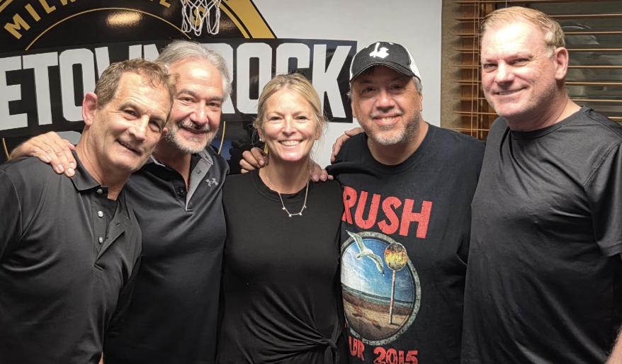 Five adults standing close together indoors, smiling at the camera with a large banner behind them. Context: group photo in front of a backdrop or sign.