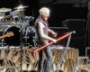 Male guitarist with bleached hair wearing a sleeveless jacket, playing a red glowing guitar on stage with a drum kit behind him.