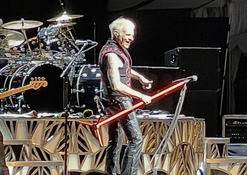 Male guitarist with bleached hair wearing a sleeveless jacket, playing a red glowing guitar on stage with a drum kit behind him.